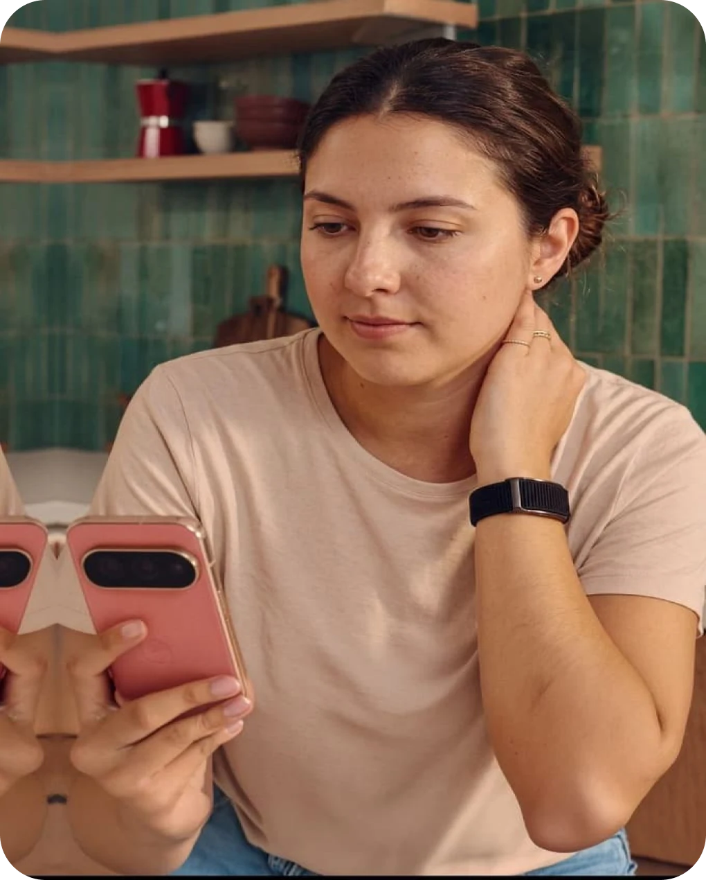 Woman in a teal-tiled kitchen checking her phone, hand at her neck, wearing the HLTH Band