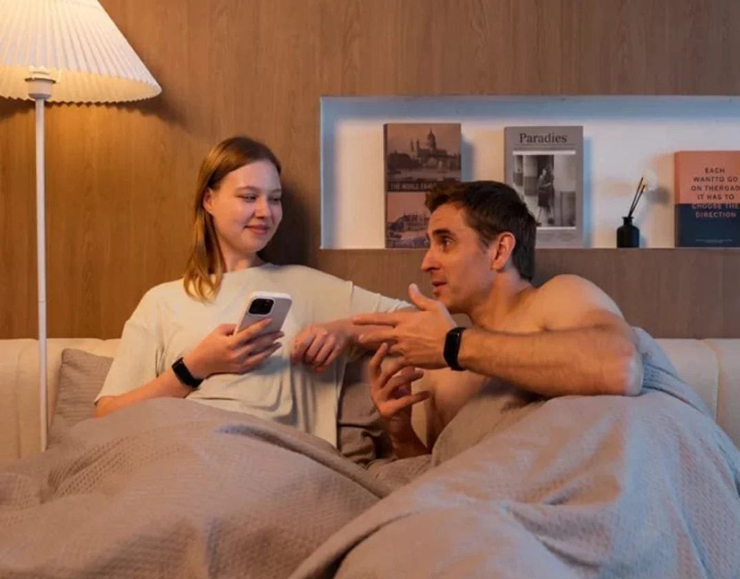 Couple in bed, one checking the HLTH app on her phone, both wearing the HLTH Band