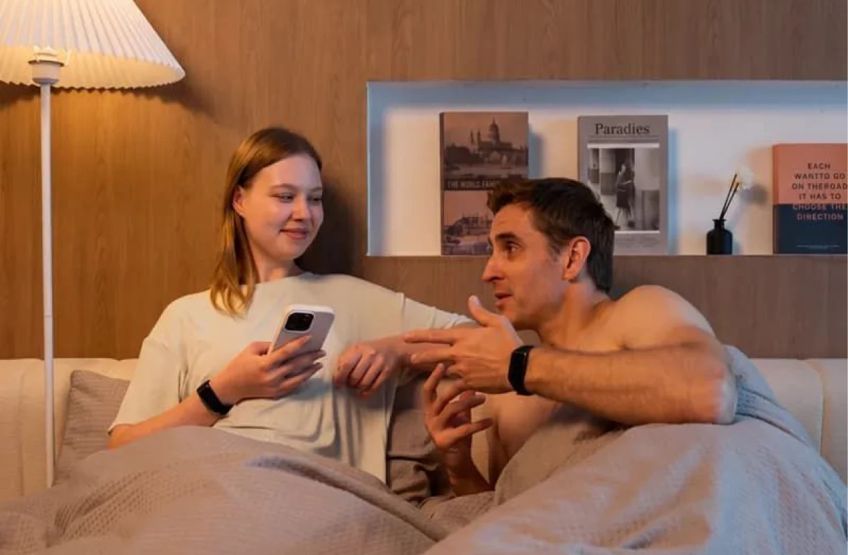 Couple in bed, one checking the HLTH app on her phone, both wearing the HLTH Band